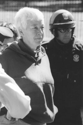 The black-and-white image displays a white-haired middle-aged woman being escorted/arrested by two policemen. One of the officers is wearing a helmet and sunglasses, and the other an officer's formal hat.