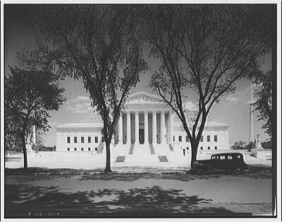 A picture of the whole U.S. Supreme Court Building from front and center. A neo-classical white building with 8 columns. Circa 1920s-1950s.